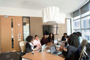 A diverse group of professionals engaged in a discussion around a modern conference table with laptops, emphasizing collaboration and inclusivity in workplaces for wellness.