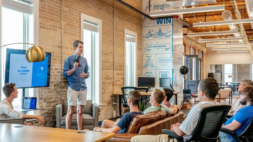 A man presents to a group of colleagues in a modern office with exposed brick walls and large windows, showcasing a collaborative environment that fosters workplaces for wellness.