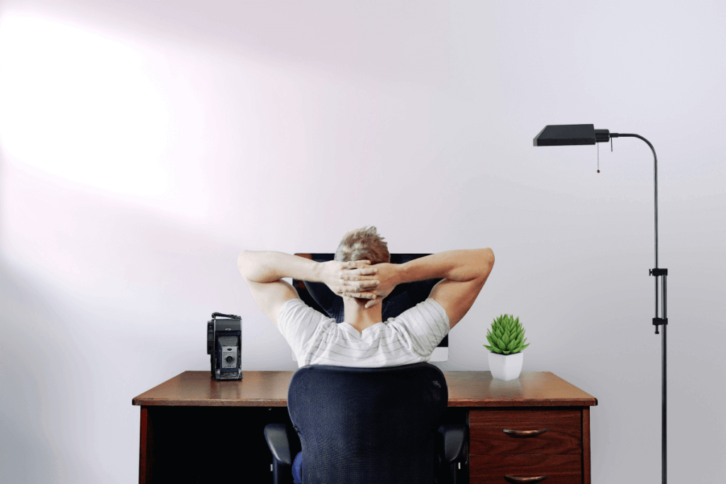 A man sits in an ergonomic office chair, leaning back with his hands behind his head, demonstrating a relaxed posture. His workspace features a well-placed computer screen, a vintage camera, a potted plant, and a floor lamp, highlighting an ergonomic and organized setup.