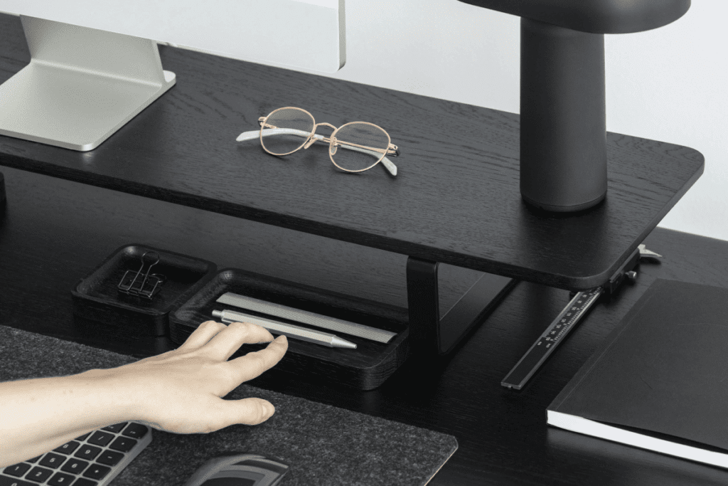 A minimalist black office desk with a monitor stand, ergonomic accessories, a black lamp, and neatly organized stationery, creating a stylish and functional workspace.