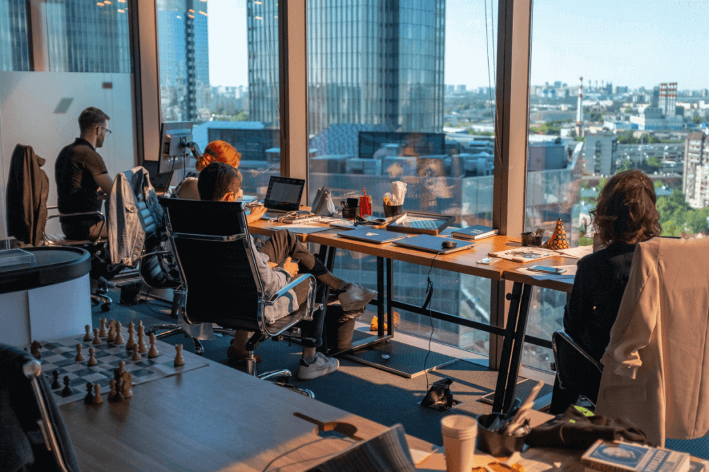 Modern office with professionals engaged in remote work at a shared desk, large windows overlooking the city skyline, and a chessboard in the foreground.