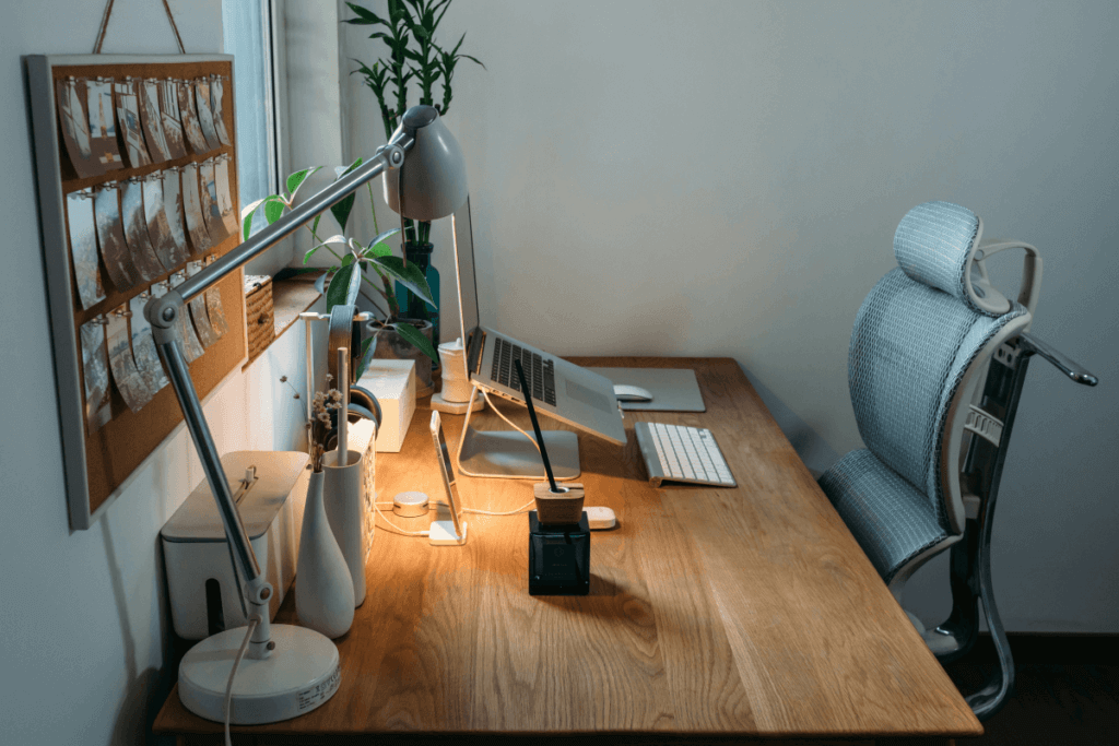 A modern office desk setup with a wooden surface, an ergonomic mesh chair, a laptop on a stand, a desk lamp, and decorative elements like plants and a corkboard with pinned photos. The workspace emphasizes style, organization, and a cozy atmosphere.