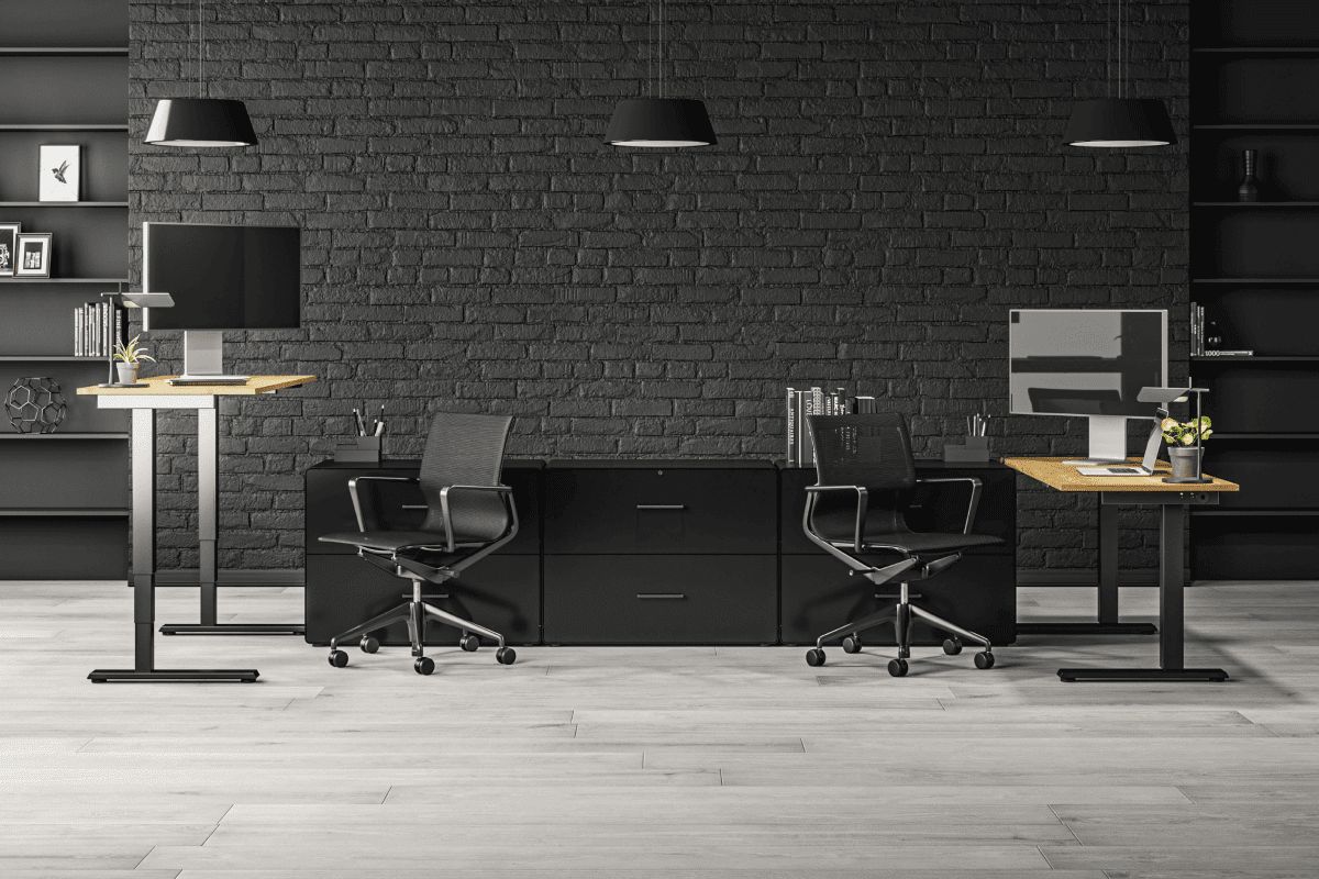 Modern office setup with black ergonomic chairs, adjustable standing desks with wooden surfaces, and dual monitors against a dark brick wall