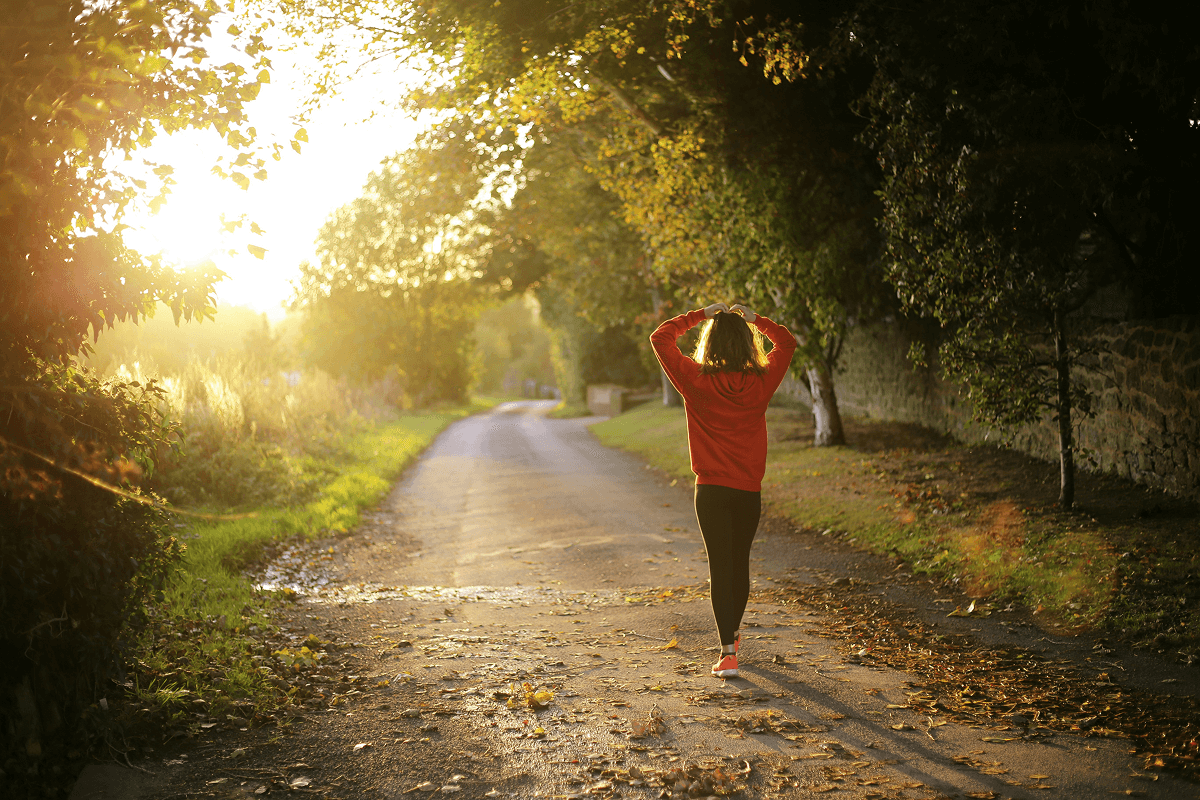 A woman in a red sweatshirt walks on a sunlit country road surrounded by trees and fallen autumn leaves during golden hour, symbolizing the benefits of work-life balance and the importance of taking time for relaxation and reflection.