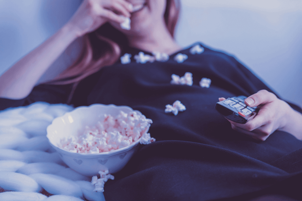 Woman relaxing after remote work, watching TV while eating popcorn, holding a remote control, and lying on a couch.