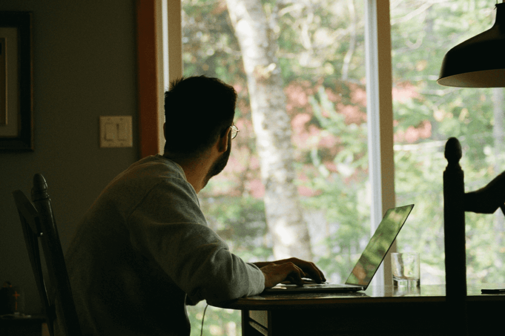 Man working from home on a laptop at a wooden desk, looking out a large window with trees in the background.
