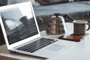 A MacBook Air displaying code on a white table, accompanied by a coffee mug, a stainless steel kettle, and a smartphone, creating a remote work setup.