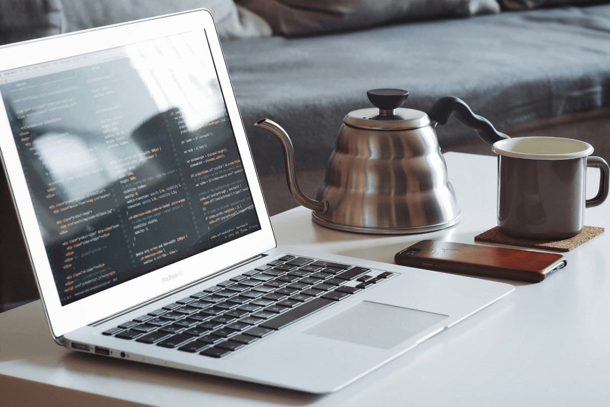 A MacBook Air displaying code on a white table, accompanied by a coffee mug, a stainless steel kettle, and a smartphone, creating a remote work setup.