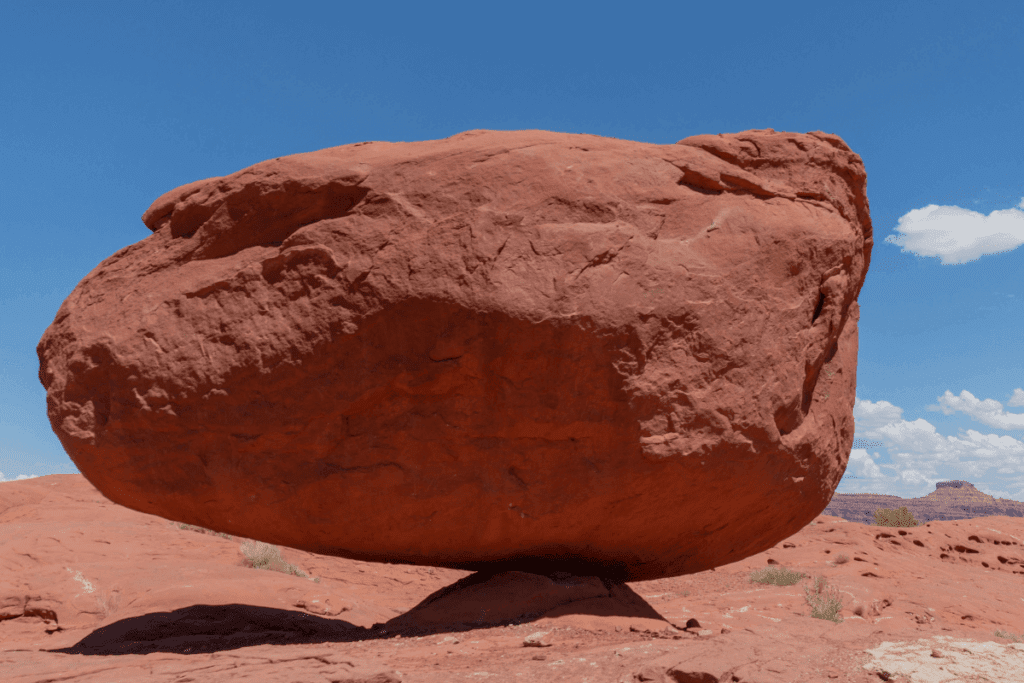 A large red sandstone rock balancing on a small pedestal in a desert landscape under a clear blue sky.