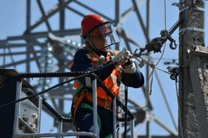 An electrician in a red hard hat and orange safety vest works on a utility pole, handling electrical equipment, demonstrating skills essential for how to become an electrician.