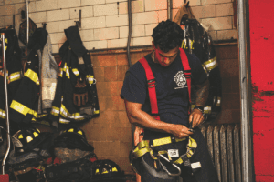 A firefighter adjusts their gear in a station surrounded by jackets and helmets on the wall, illustrating the preparation process and how to become a volunteer firefighter by understanding equipment readiness.
