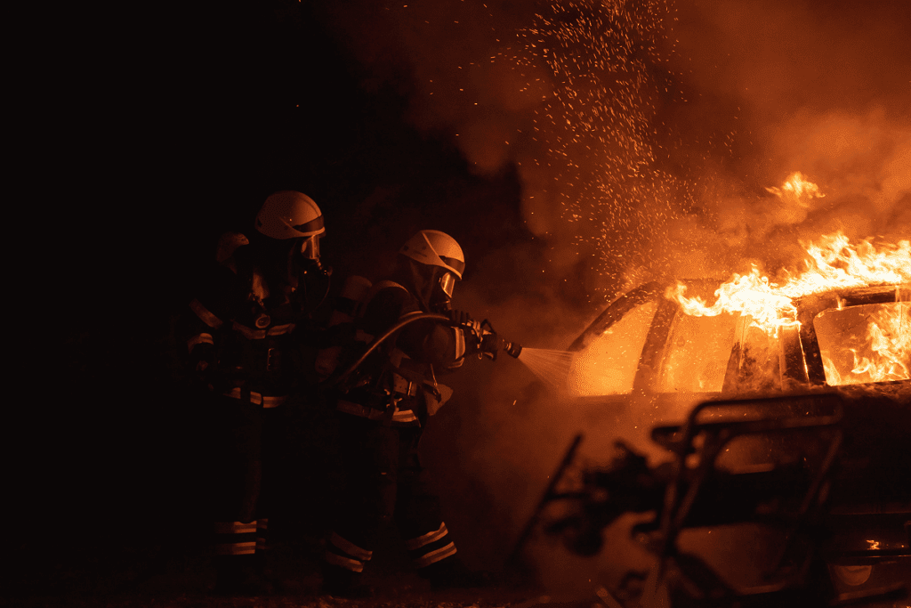 Two firefighters in gear use a hose to extinguish a vehicle fire, with flames and smoke rising, demonstrating teamwork in action and how to become a volunteer firefighter by learning to handle emergencies like this.