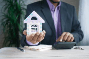 A business professional in a suit holding a pastel-colored model house at a desk with a calculator, representing real estate management, ideal for those exploring how to become a property manager.