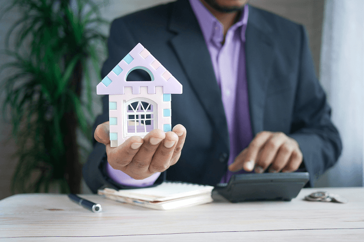 A business professional in a suit holding a pastel-colored model house at a desk with a calculator, representing real estate management, ideal for those exploring how to become a property manager.