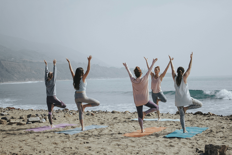 A group of people practice yoga on a beach, standing on mats in tree pose with arms raised, facing the ocean with waves and cliffs in the background, showcasing how to become a yogi in a serene outdoor setting.