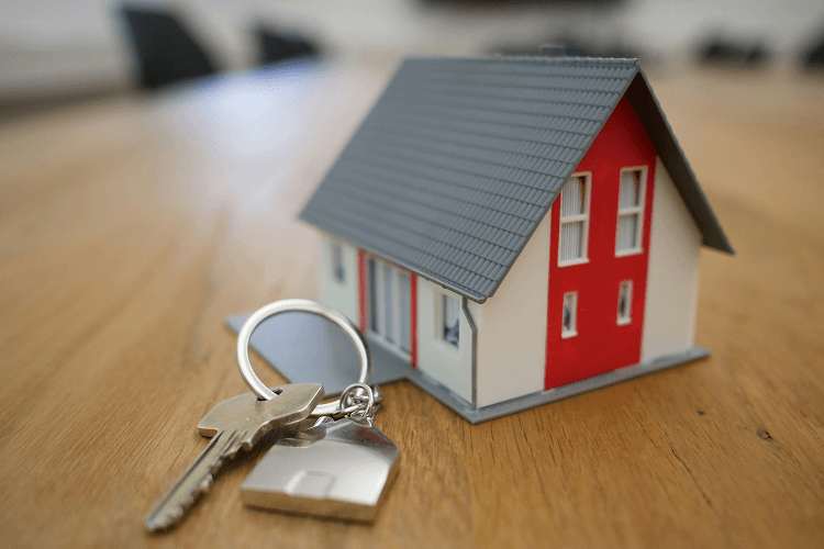 A small model house with red accents and a key on a keyring, placed on a wooden surface, illustrating property ownership and management, useful for understanding how to become a property manager.