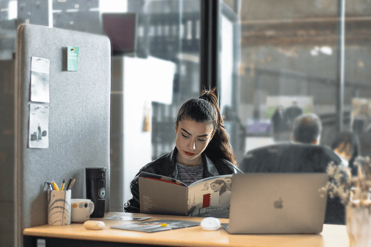 A person reading a book at a modern office desk with a laptop and coffee mug, multitasking with 2 jobs at the same time. Office supplies and a blurred background enhance the work environment.