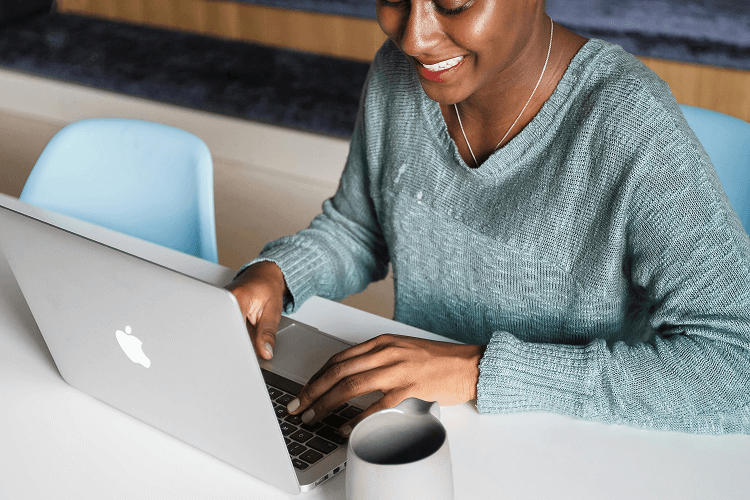 A person typing on a laptop at a table with a coffee mug, wearing a sweater, efficiently managing 2 jobs at the same time. The blurred office background with blue chairs suggests a productive workspace.