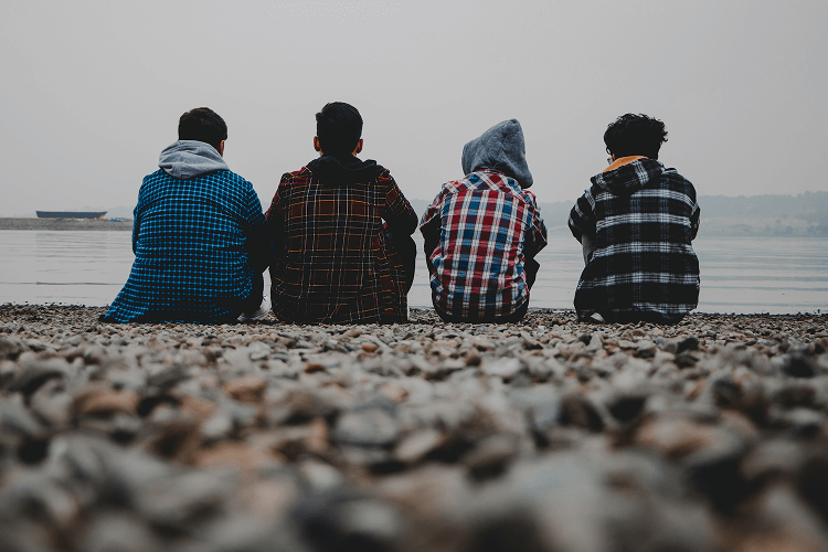 Four teens wearing flannel shirts sitting on a rocky shore looking at the water, reflecting on friendship and free time between opportunities for what jobs hire at 13.