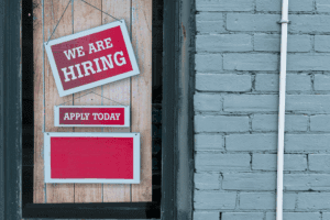 A store window featuring a red and white 'We Are Hiring' sign and an 'Apply Today' sign, offering 2 jobs at the same time. Set against a wooden background with a brick wall, ideal for job seekers.