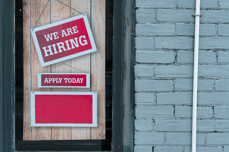 A store window featuring a red and white 'We Are Hiring' sign and an 'Apply Today' sign, offering 2 jobs at the same time. Set against a wooden background with a brick wall, ideal for job seekers.